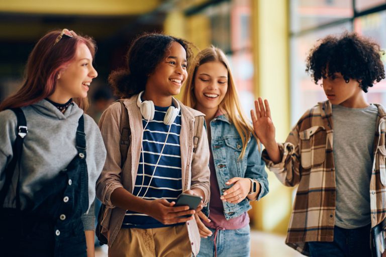 Four smiling teenagers walking together in what appears to be a school hallway. One girl in the center holds a smartphone and wears white headphones around her neck, while another girl beside her waves at the camera. The group appears happy and engaged with one another.